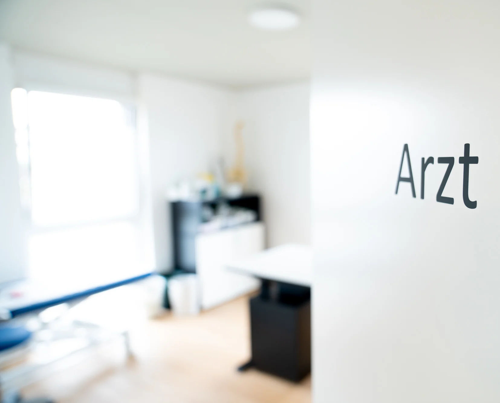 Blick durch die Praxistür mit der Aufschrift Arzt in einen hellen Behandlungsraum